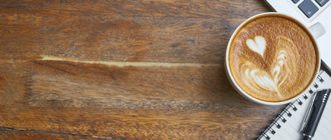 A cup of coffee on a wooden desk beside the corner of a laptop and a spiral notepad with a pen