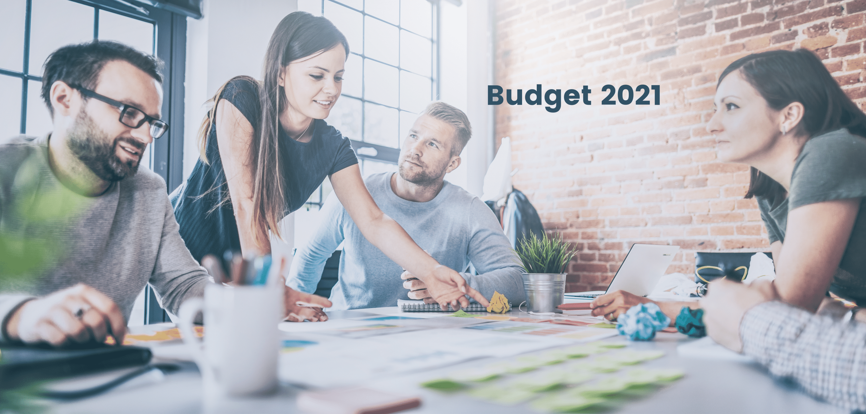 Four colleagues around a desk of printed charts and sticky notes in a brick-walled office, under the words Budget 2021.
